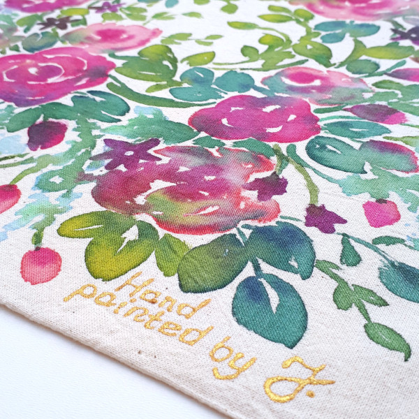 Zdjęcie przedstawia przybliżenie fragmentu torby ze złotym podpisem artystki. Torba bawełniana w kolorze ecru z ręcznie namalowanym motywem kwietnym - róże w odcieniach czerwieni i różu (w rozkwicie i w pączkach) w otoczeniu gałązek w różnych odcieniach zieleni.